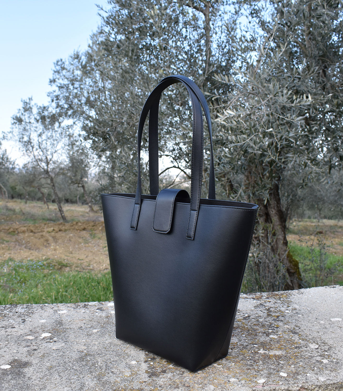Balancing the material world and the enivronment. Sustainably made in Italy. Zero-Waste Luxury. Black tote bag sitting ion cement block on n olive tree vineyard