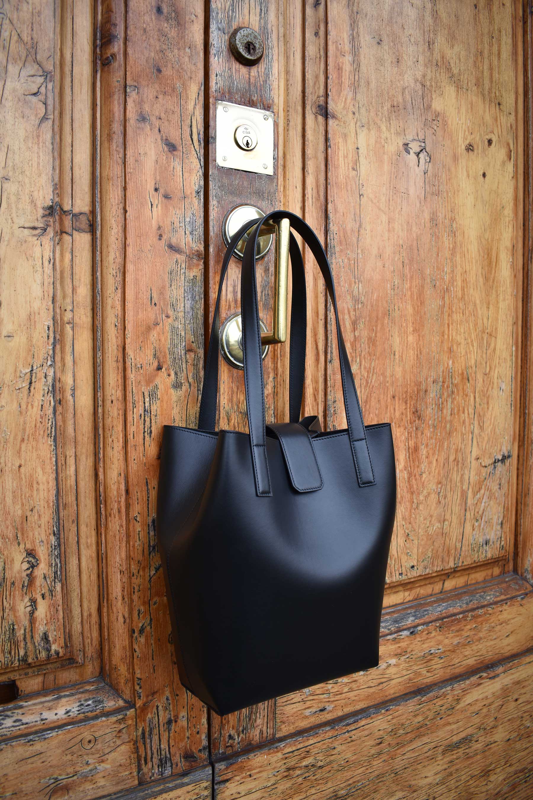 Chandra Keyser La Borsa Black Bucket Tote Bag Lightweight with two shoulder straps, snap closure, separate micro-suede zip pouch and inside clasp to contract and expand bag. Hanging on a wooden door.