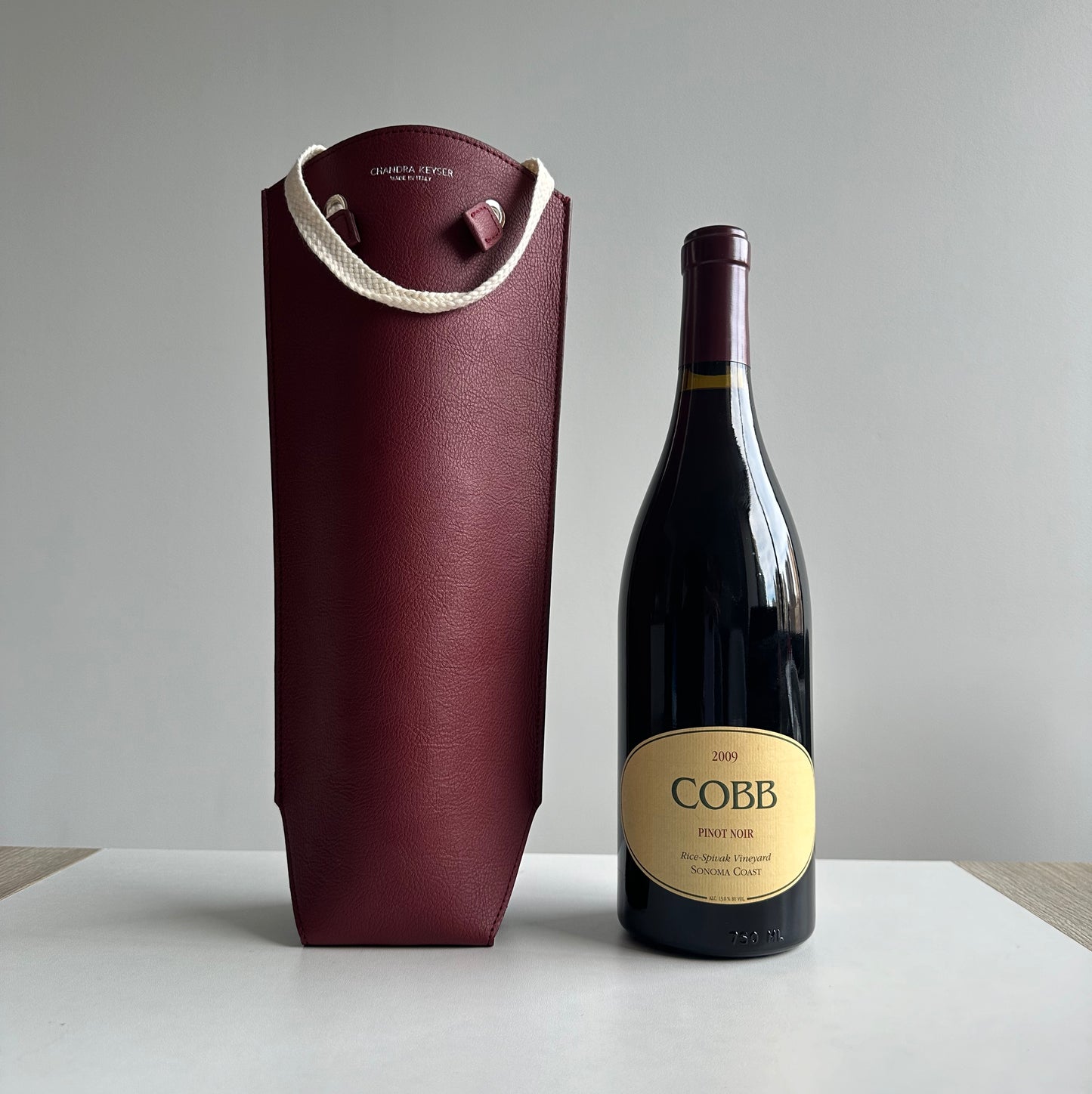 Burgendy Red Bag Wine Carrier Sustainable Luxury Gift Circular Product Supports the Environment sitting on a round table with white wall in the background and bottle of Cobb Wine 2009